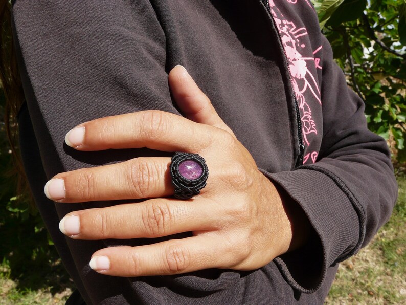 Primo pieno di anello nero con pietra preziosa ovale viola Rubino, indossato da donna con felpa nera, su sfondo verde alla luce del sole.