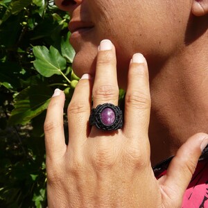 Primo pieno di anello nero, con al centro grande pietra preziosa naturale viola Rubino Stella, indossato da donna con mano su volto, su sfondo verde alla luce del sole.