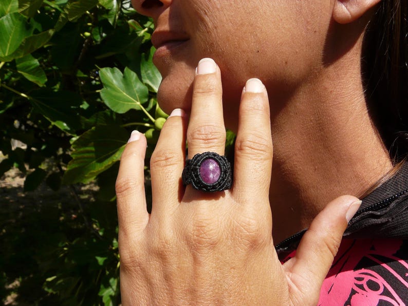 Primo pieno di anello nero, con al centro grande pietra preziosa naturale viola Rubino Stella, indossato da donna con mano su volto, su sfondo verde alla luce del sole.