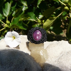 Könnte beinhalten: Handgefertigter Makramee-Ring mit einem großen, ovalen, lila Edelstein, gefasst in einem schwarzen gewebten Band. Der Ring wird auf einer weißen Oberfläche mit einer weißen Blume und grünem Laub im Hintergrund präsentiert. Ein einzigartiger Schmuck.