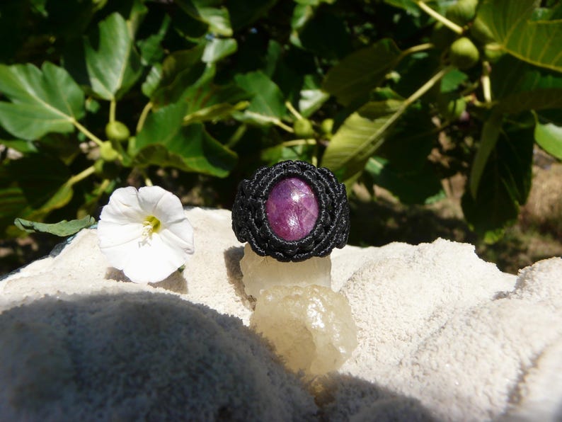 Primo piano di anello nero fatto a mano in Macramè con al centro una pietra preziosa naturale viola Rubino, con asterismo, di forma ovale e taglio lucido. Gioiello olistico esposto su minerale grezzo bianco con fiore bianco su sfondo a foglie verdi.