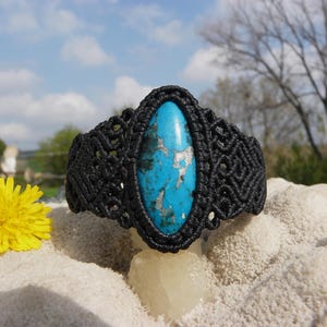 May include: A black macrame bracelet with a large, oval turquoise stone. The bracelet is displayed on a light-coloured surface with a yellow dandelion flower in the foreground. The background shows a blue sky and trees.