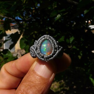 Può includere: Un anello fatto a mano con una pietra preziosa di opale a forma di pera, incastonata in un design macramè grigio. L'opale mostra un vibrante gioco di colori, con lampi di verde, blu e rosso. L'anello è tenuto su uno sfondo verde sfocato.