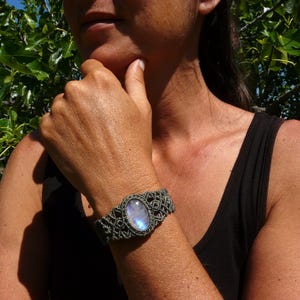 May include: A handmade macrame bracelet with a large, oval moonstone. The bracelet is gray and features intricate knotting around the central stone. The moonstone has a blue and white iridescent glow. The bracelet is worn on a wrist.
