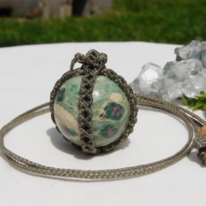 May include: A green and beige stone pendant necklace. The stone is round with red and green markings, encased in a woven, gray-green macrame. The necklace cord is a matching gray-green color, with a wooden bead accent.