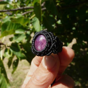 Primo piano laterale sinistro di anello nero, con al centro grande pietra preziosa naturale viola Rubino Stella, tenuto in mano su sfondo verde natura, alla luce del sole.