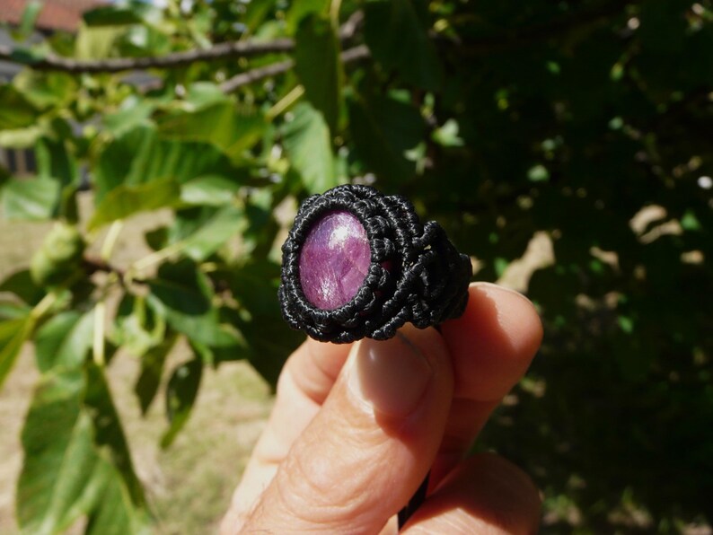 Primo piano laterale sinistro di anello nero, con al centro grande pietra preziosa naturale viola Rubino Stella, tenuto in mano su sfondo verde natura, alla luce del sole.