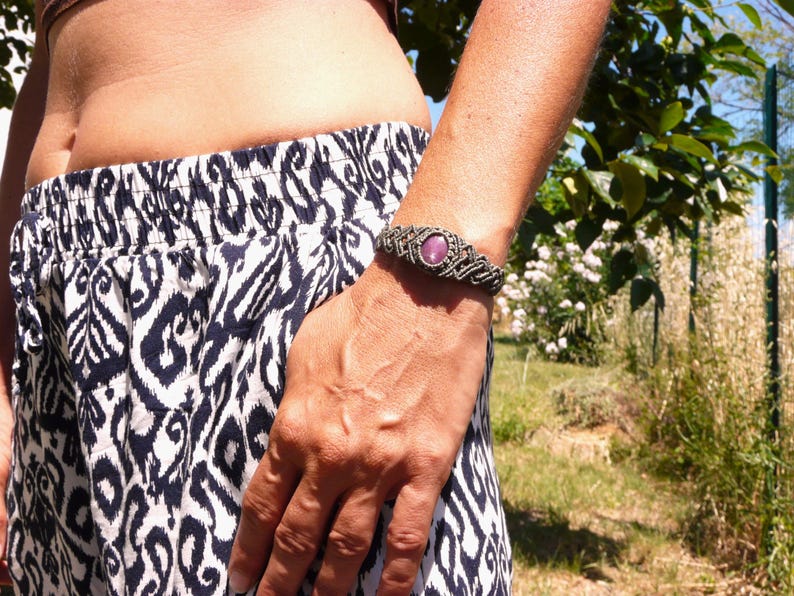 Bracciale a manetta intrecciato a mano in Macramè filato color grigio-verde con pietra naturale Rubino Stella, indossato da ragazza con pantaloni bianche e neri su sfondo prato verde.