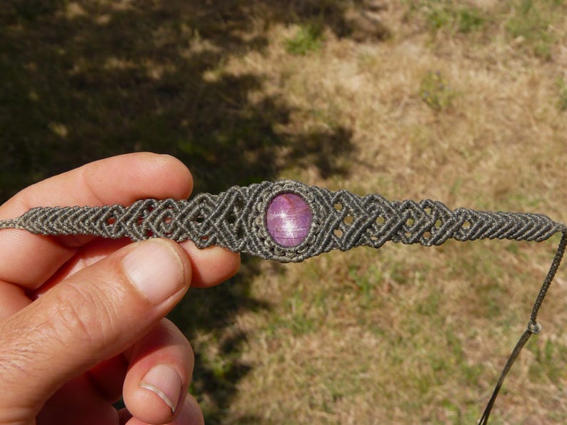Bracciale a manetta intrecciato a mano in Macramè filato color grigio-verde con pietra naturale Rubino Stella vista bracciale aperto con dettaglio sull'intreccio geometrico in Macramè, primo piano su sfondo prato verde.