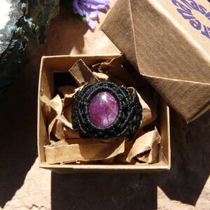 Primo piano di anello nero intrecciato in Macramè, con al centro grande pietra preziosa naturale viola Rubino Stella, esposto in confezione regalo con logo Rasikoserena alla luce del sole.