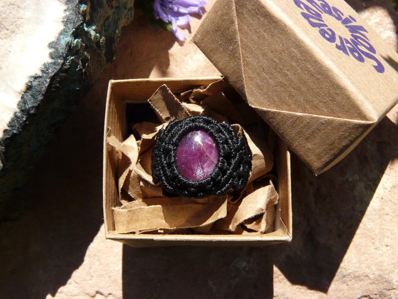 Primo piano di anello nero intrecciato in Macramè, con al centro grande pietra preziosa naturale viola Rubino Stella, esposto in confezione regalo con logo Rasikoserena alla luce del sole.