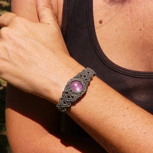 Bracciale a manetta intrecciato a mano in Macramè filato color grigio-verde con pietra naturale Rubino Stella, indossato da ragazza su sfondo naturale.