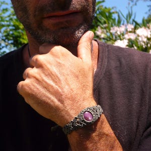 Bracciale a manetta intrecciato a mano in Macramè filato color grigio-verde con pietra naturale Rubino Stella primo piano indossato da uomo con maglia nera sullo sfondo fiori di Oleandro.