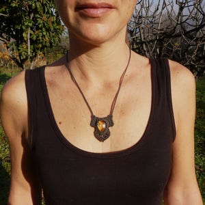 May include: A necklace featuring a teardrop-shaped tiger's eye gemstone set in a dark brown macrame cord pendant. The intricate knotted cord design surrounds the stone, which displays golden-brown chatoyancy, suspended from a thin cord necklace.