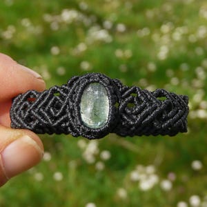Puede incluir: Un brazalete de macramé negro con una piedra preciosa ovalada de color verde claro. El brazalete se muestra sobre un fondo borroso verde y blanco. El diseño de macramé es detallado e intrincado.