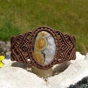 Può includere: Un bracciale in macramè marrone con una pietra di forma ovale al centro. La pietra presenta una base bianca e traslucida con tre distinte inclusioni arrotondate color marrone dorato disposte verticalmente al centro della superficie lucida.