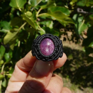 Primo piano di anello nero, con al centro grande pietra preziosa naturale viola Rubino Stella, tenuto in mano su sfondo verde natura, alla luce del sole.