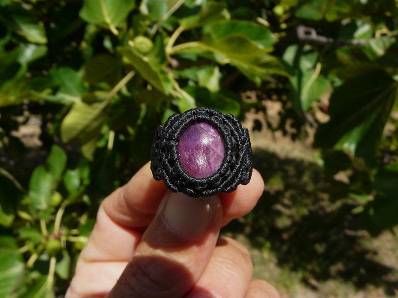 Primo piano di anello nero, con al centro grande pietra preziosa naturale viola Rubino Stella, tenuto in mano su sfondo verde natura, alla luce del sole.