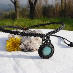 Può includere: Collana in macramè nera con un ciondolo rotondo in pietra azzurra. La collana è esposta su una superficie bianca con un fiore giallo e un gruppo di cristalli. La collana è regolabile.
