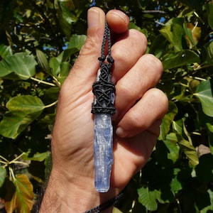 Puede incluir: Colgante de cristal de cianita azul con cordón negro tejido. El cristal se sostiene en una mano sobre un fondo de follaje verde. El colgante presenta un diseño detallado de nudos negros en la parte superior.