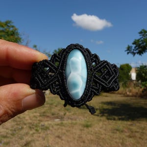 May include: A bracelet featuring a central oval-shaped light blue and white Larimar gemstone set within a black macrame cord band. The cord is intricately knotted in a geometric pattern surrounding the stone, with two small dangling cord ends.