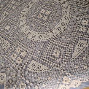Puede incluir: Un mantel de crochet blanco con un diseño circular en el centro. El mantel tiene un delicado patrón floral y un borde festoneado.