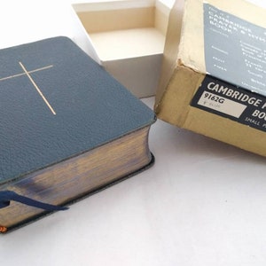 Pode incluir: Um livro de orações de Cambridge encadernado em couro azul com uma cruz dourada na capa. O livro está em uma caixa de papelão com o texto "Cambridge Prayer Book" impresso nela.