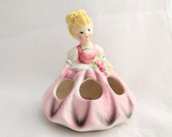 Może przedstawiać: Ceramiczna figurka dziewczyny w różowej sukience z bukietem kwiatów. Sukienka ma pięć otworów na przechowywanie szminek.