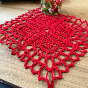 Può includere: Un centrino all'uncinetto rosso con un delicato motivo floreale. Il centrino è di forma quadrata e ha un foro al centro.