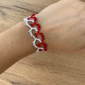 Op de afbeelding: Een witte en rode kralen armband met een delicaat, ingewikkeld ontwerp. De armband is gemaakt met kleine, ronde kralen en heeft een herhalend patroon van lussen en bogen.