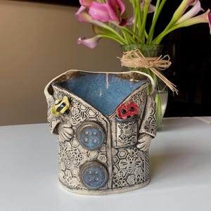 May include: A ceramic vase shaped like a jacket, with a blue interior. The jacket is decorated with embossed patterns, blue buttons, and a pocket with red flowers. A yellow butterfly is on the lapel. A vase of pink calla lilies is in the background.