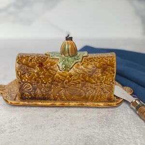 May include: A ceramic butter dish shaped like a loaf of bread with a leafy design and a pumpkin-shaped lid. The dish is brown and green with a matching knife.