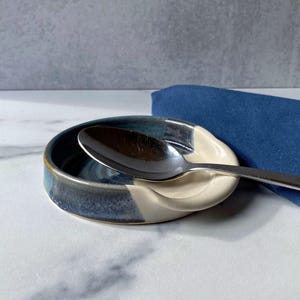 May include: A blue and white ceramic spoon rest with a spoon resting inside. The spoon rest has a wavy edge and a smooth, white interior.