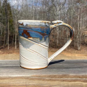 May include: A handmade ceramic mug with a unique design. The mug features a white base with diagonal lines and a blue and brown glazed top. The handle is curved and the mug is sitting on a wooden surface.