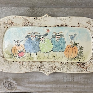 May include: A white ceramic serving tray with a decorative floral border. The tray features a hand-painted scene of three sheep with pumpkins and a bluebird.