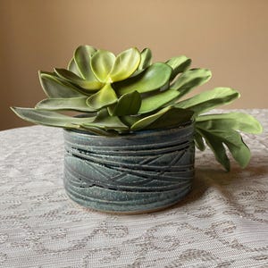 May include: A cylindrical ceramic planter with a textured, dark blue-green glaze featuring horizontal carved lines. The planter holds a large, artificial succulent plant with thick, fleshy, light green leaves that fan out over the rim.