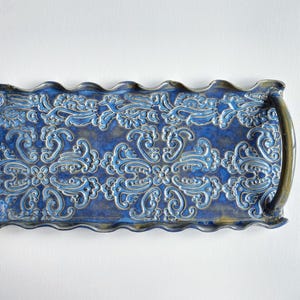 May include: A rectangular ceramic tray with a blue and gold glaze. The tray features a repeating floral pattern in a lighter blue, with a scalloped edge. The tray is approximately 12 inches long and 6 inches wide.