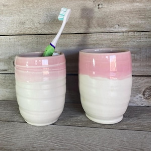 May include: Two ceramic bathroom tumblers with a pink and white glaze. The tumblers are cylindrical and have a slightly rounded bottom. One tumbler has a toothbrush inside.