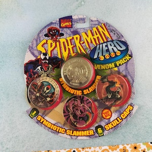 Könnte beinhalten: Ein Spider-Man Hero Venom Pack mit Symbiotic Slammer und Skull Caps. Die Verpackung zeigt Spider-Man- und Venom-Charaktere. Das Paket enthält eine Silbermünze und mehrere farbige Kappen mit Charakterdesigns.