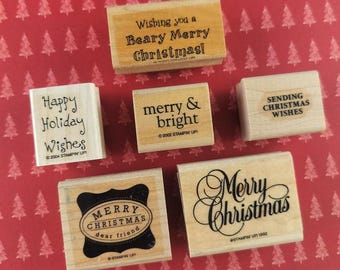 以下が含まれることがあります： 黒いインクで様々なクリスマスのメッセージが刻印された6つの長方形の木製ラバースタンプ。スタンプには「Wishing you a Beary Merry Christmas」、「Happy Holiday Wishes」、「merry & bright」、「SENDING CHRISTMAS WISHES」、「MERRY CHRISTMAS dear friend」、「Merry Christmas」というフレーズが含まれています。