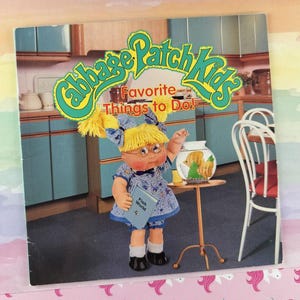 May include: A Cabbage Patch Kids book titled Favorite Things to Do! featuring a blonde doll with pigtails wearing a blue floral dress and glasses, standing next to a small table with a fishbowl in a kitchen setting with blue cabinets.