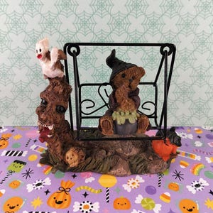 May include: A Halloween-themed figurine featuring a brown teddy bear wearing a black witch hat and sitting on a black metal swing. A white ghost sits on a brown tree stump next to the swing, which also includes a small skull and a pumpkin.