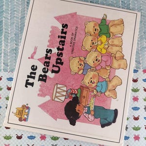 以下が含まれることがあります： 「The Bears Upstairs」というタイトルの子供向けの本。テディベアと女の子のイラストが描かれています。表紙にはピンク色の城のデザインと「A Book of Creative Dramatics」という文字が描かれています。本は模様のある表面にあります。