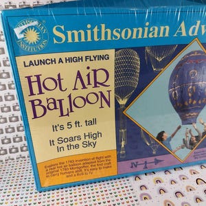 Op de afbeelding: Een Smithsonian Adventures heteluchtballon kit. De blauwe doos heeft de tekst "Lanceer een hoogvliegende heteluchtballon". De kit is 1,52 meter hoog en stijgt hoog in de lucht.