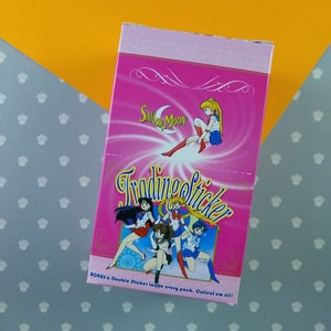 May include: A pink box containing a Sailor Moon trading sticker pack. The box features a Sailor Moon character with a pink background and the text "Sailor Moon Trading Sticker" in white. The text "Bonus - a Double Sticker inside every pack. Collect'em all!" is printed on the bottom of the box.