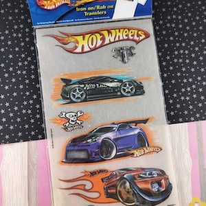 May include: Sheet of Hot Wheels iron-on transfers featuring various car designs and logos. The sheet includes images of race cars in black, purple, and orange, with the Hot Wheels logo and text. The packaging reads "Iron on/Rub on Transfers."