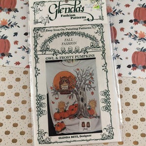 Pode incluir: Um pacote de Glenda's Fashion Patterns com um desenho para passar a ferro. O desenho inclui uma coruja, abóboras e uma árvore, com o texto "FALL FASHION" e "OWL & FROSTY PUMPKINS". O pacote está em um fundo padronizado.
