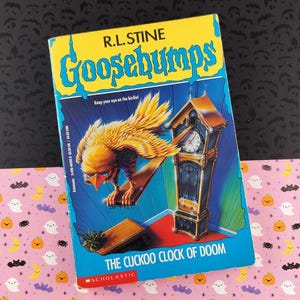May include: A vintage paperback book titled "Goosebumps: The Cuckoo Clock of Doom" by R.L. Stine. The cover features a yellow and blue design with a menacing bird and a grandfather clock. The book is set against a patterned background.