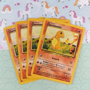 May include: Several Pokémon trading cards featuring the character Charmander. The cards have a yellow border and include the character's name, HP, and attack details. The background is a light blue pattern with unicorns and rainbows.
