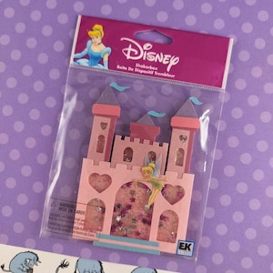 May include: A Disney shakerbox featuring a pink castle design with Tinkerbell and heart cutouts. The packaging includes the Disney logo, the text "Shakerbox" and "Boite De Dispositif Trembleur". The shakerbox is in a clear plastic package.